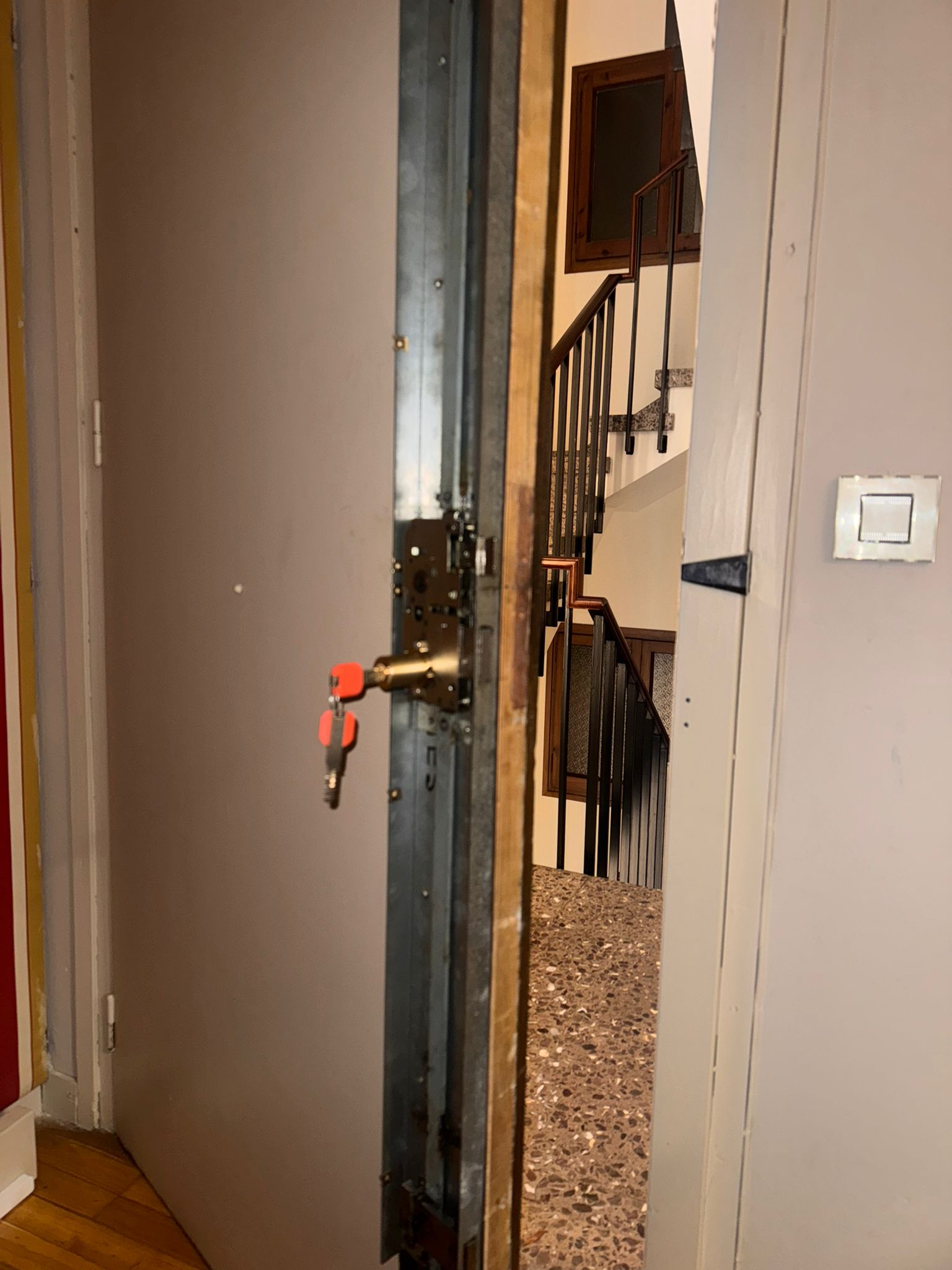 Puerta acorazada abierta con cerradura multipunto y llaves en bombín, con escalera comunitaria visible al fondo
